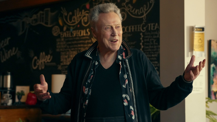 Christopher Walken smiles, gesturing with his arms, indoors