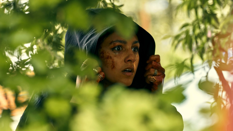 Rhianne Barreto is hidden amongst leaves, gripping her hood, face covered in a small amount of blood