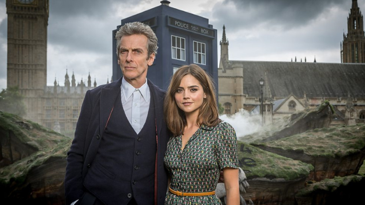Jenna Coleman and Peter Capaldi stand side-by-side, in character as Clara Oswald and the Doctor, in front of the TARDIS, outdoors in London 