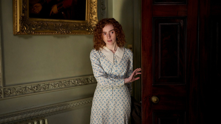 Eildh Fisher as Evie Galloway, stood by an open door in a grand-looking house, peering into the room