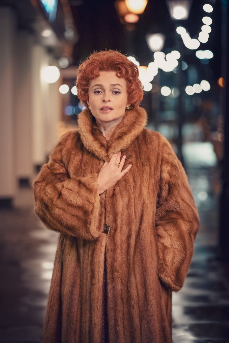 A glamorous woman with auburn hair and a tan fur coat stands outside at night in front of street lamps