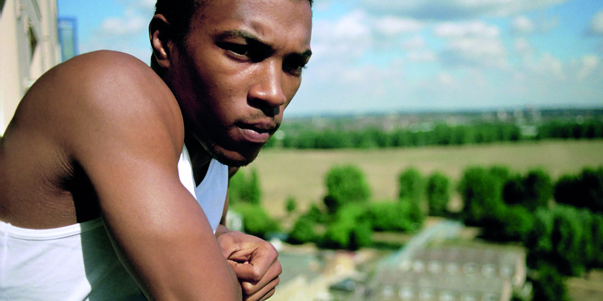 A young Jamaican boy looks into the distance to the right of the camera, he is in front of an expanse of fields and trees. He wears a white tank top and has facial hair beginning to grow.